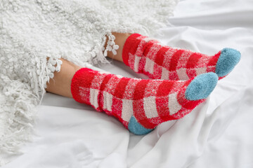 Female legs in knitted socks covered with plaid on bed, closeup. Concept of heating season