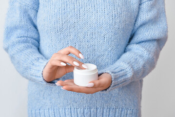 Woman in warm sweater holding jar with cream on light background, closeup