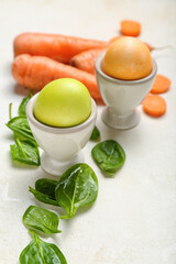 Holders with beautiful Easter eggs, carrot and spinach leaves on light background, closeup