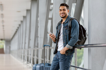 Portrait Of Cheerful Young Arab Guy Using Smartphone While Waiting At Airport