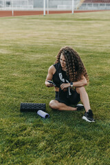 Woman athlete is sitting on a lawn at a stadium and typing in her phone.