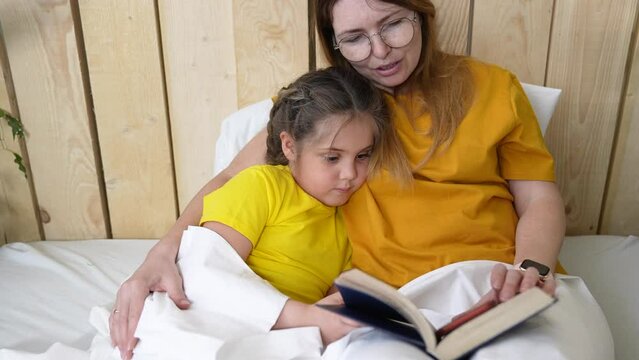 mother in bed reading a book to her daughter. mother lifestyle and child girl reading a book in bed before going to sleep. happy family kid dream concept indoor