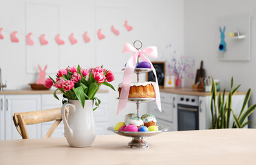Stand with Easter cakes, eggs and tulips on dining table in kitchen