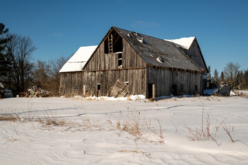 An aging wooden pole barn slowly collapsing.  Shot on a sunny day in winter with fresh snow and blue sky.