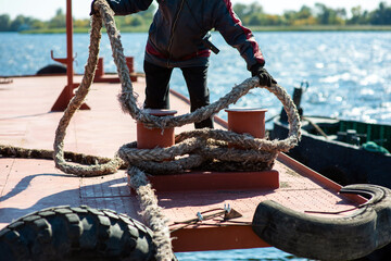 Sailor in overalls on the deck of the ship pulls the mooring cable in the port - obrazy, fototapety, plakaty