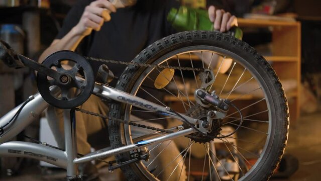 A slender guy repairs a broken bike in the garage. A man works in a room with old bicycle equipment. Repair of equipment with your own hands. The guy lubricates the chain and wheels with oil