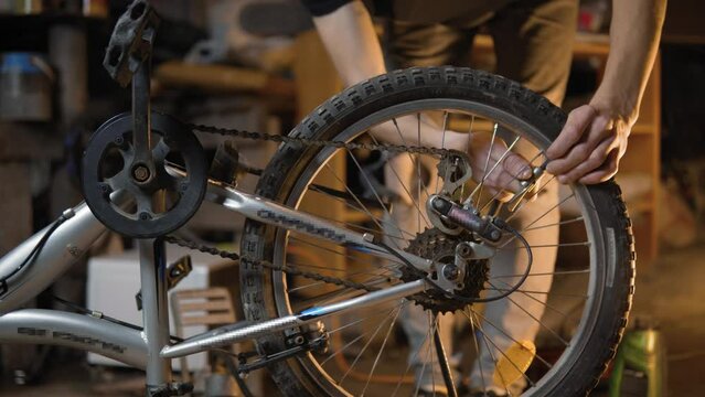 A slender guy repairs a broken bike in the garage. A man works in a room with old bicycle equipment. Repair of equipment with your own hands. The guy lubricates the chain and wheels with oil