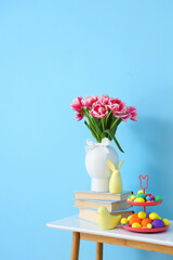 Vase with tulips, books, decor and Easter eggs on table near blue wall