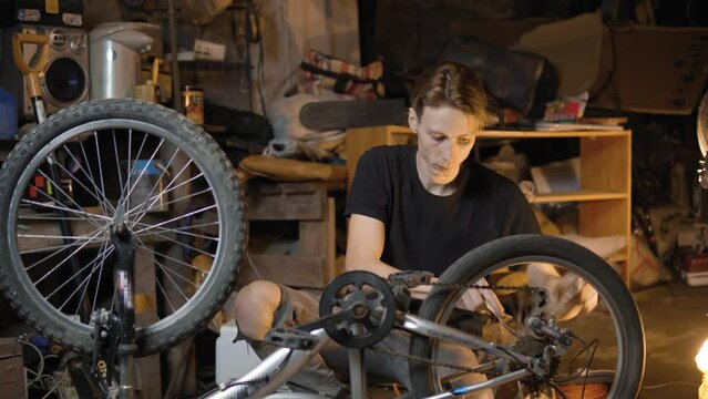 A slender guy repairs a broken bike in the garage. A man works in a room with old bicycle equipment. Repair of equipment with your own hands. The guy lubricates the chain and wheels with oil