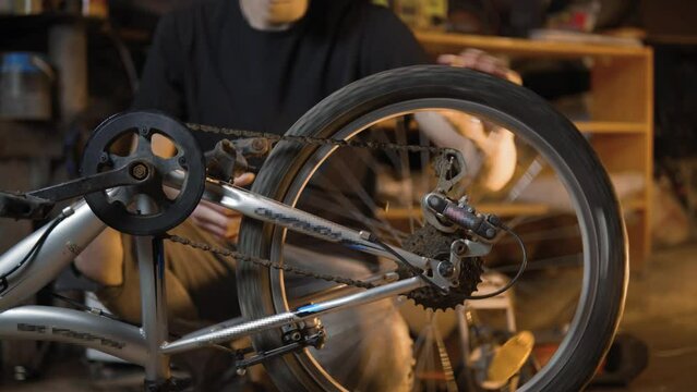 A slender guy repairs a broken bike in the garage. A man works in a room with old bicycle equipment. Repair of equipment with your own hands. The guy lubricates the chain and wheels with oil