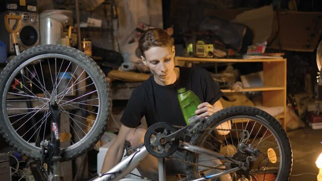 A slender guy repairs a broken bike in the garage. A man works in a room with old bicycle equipment. Repair of equipment with your own hands. The guy lubricates the chain and wheels with oil