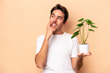 Young caucasian man holding a plant isolated on beige background relaxed thinking about something looking at a copy space.