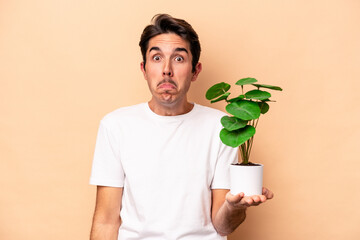 Young caucasian man holding a plant isolated on beige background shrugs shoulders and open eyes...