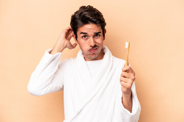 Young caucasian man wearing a bathrobe holding a toothbrush isolated on beige background being...