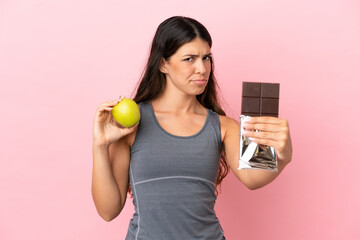 Young caucasian woman isolated on pink background taking a chocolate tablet in one hand and an...