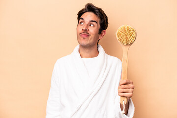 Young caucasian man wearing a bathrobe holding a back scratcher isolated on beige background dreaming of achieving goals and purposes