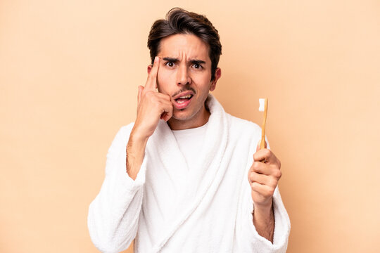 Young caucasian man wearing a bathrobe holding a toothbrush isolated on beige background showing a disappointment gesture with forefinger.