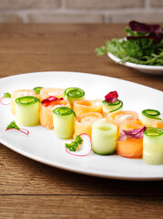 Appetizer of lightly salted salmon with tender cheese and cucumber on a white plate on a wood background. Beautiful appetizer for a festive party. Rolls of red fish and cucumber. Copy space. Close up.