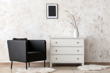 Tree branches and Easter eggs in vase on chest of drawers and armchair near light wall