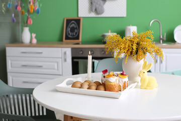 Tray with Easter cakes, eggs, burning candle and mimosa flowers on dining table in kitchen