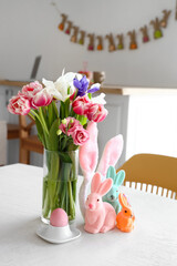 Vase with beautiful flowers, Easter rabbits and holder with painted egg on dining table in kitchen
