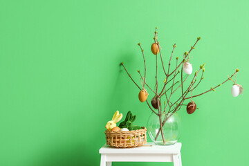 Basket with Easter eggs, rabbits and vase with tree branches on stepladder stool near color wall
