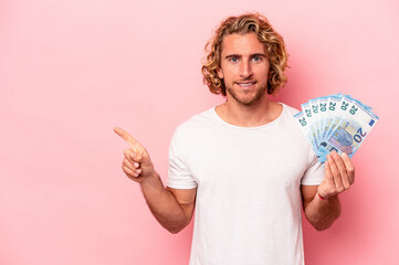 Young caucasian man holding banknotes isolated on pink background smiling and pointing aside,...