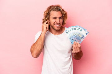 Young caucasian man holding banknotes isolated on pink background covering ears with hands.