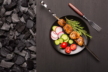 Black plate with kebab and fresh vegetables on a black wooden tablertop on charcoal for barbecue. Top view.