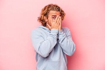 Young caucasian man isolated on pink background blink through fingers frightened and nervous.