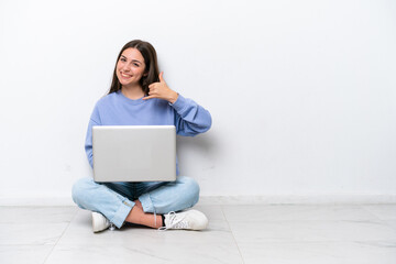 Young caucasian woman with laptop sitting on the floor isolated on white background making phone gesture. Call me back sign