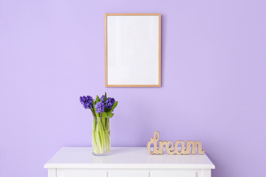 Vase with beautiful flowers, word DREAM on table and blank photo frame on color wall