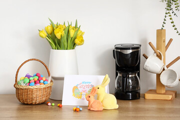 Basket with Easter eggs, rabbits, greeting card and vase with tulips on counter near light wall