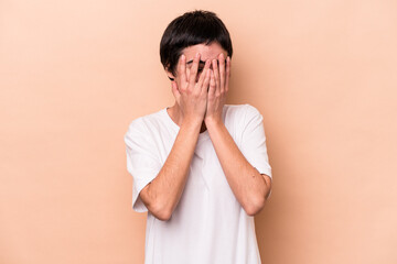 Young caucasian woman isolated on beige background blink at the camera through fingers, embarrassed covering face.