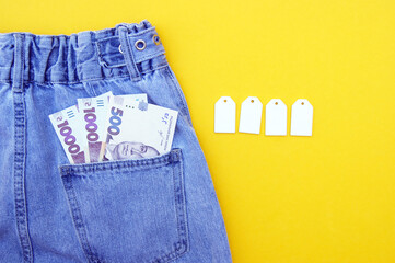Flat lay, detail of denim jeans with Ukrainian hryvnia bills in pocket with white label on yellow...
