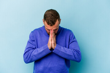 Young caucasian man isolated on blue background praying, showing devotion, religious person looking for divine inspiration.