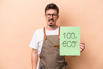 Young caucasian gardener man holding 100% eco placard isolated on beige background screaming very...