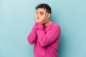 Young caucasian man isolated on blue background blink through fingers frightened and nervous.