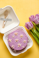 Plastic lunch box with tasty bento cake and flowers on orange background