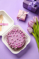 Plastic lunch box with tasty bento cake and flowers on purple background