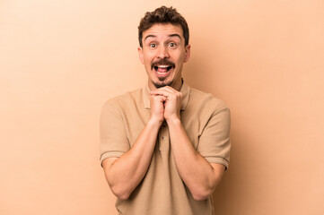 Young caucasian man isolated on beige background praying for luck, amazed and opening mouth looking to front.