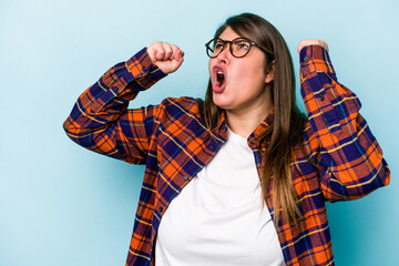 Young caucasian overweight woman isolated on blue background raising fist after a victory, winner concept.