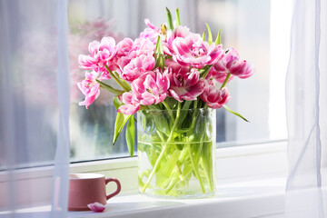 Glass vase with beautiful tulips and cup of tea on windowsill