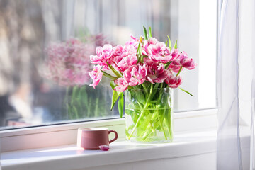 Vase with bouquet tulip flowers and cup of tea on windowsill