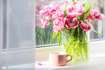 Glass vase with beautiful tulip flowers and cup of tea on windowsill