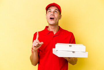 Young delivery caucasian man holding pizzas isolated on yellow background pointing upside with opened mouth.