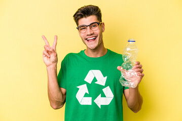 Young caucasian man holding a bottle of plastic to recycle isolated on yellow background joyful and...