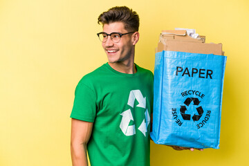 Young caucasian man holding a recycling bag full of paper to recycle isolated on yellow background looks aside smiling, cheerful and pleasant.
