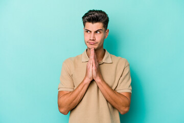 Young caucasian man isolated on blue background praying, showing devotion, religious person looking...