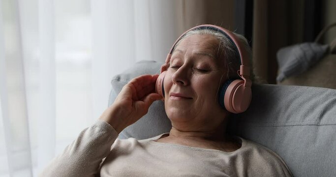 80s grey-haired woman enjoy quality sound, listen favourite music through headphones with eyes closed, relax leaned on cozy sofa at home, close up. No stress, hobby, retired life, modern tech concept